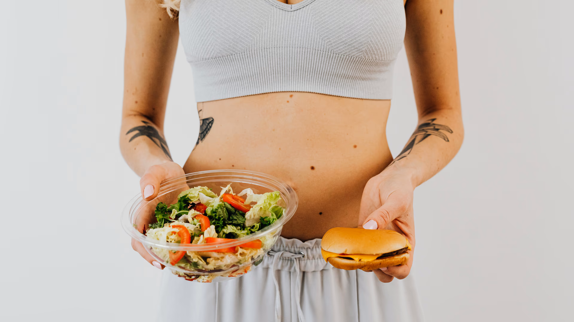 A woman holding a salad and cheeseburger, representing healthy vs junk food choices - Image by kaboompics from Pexels