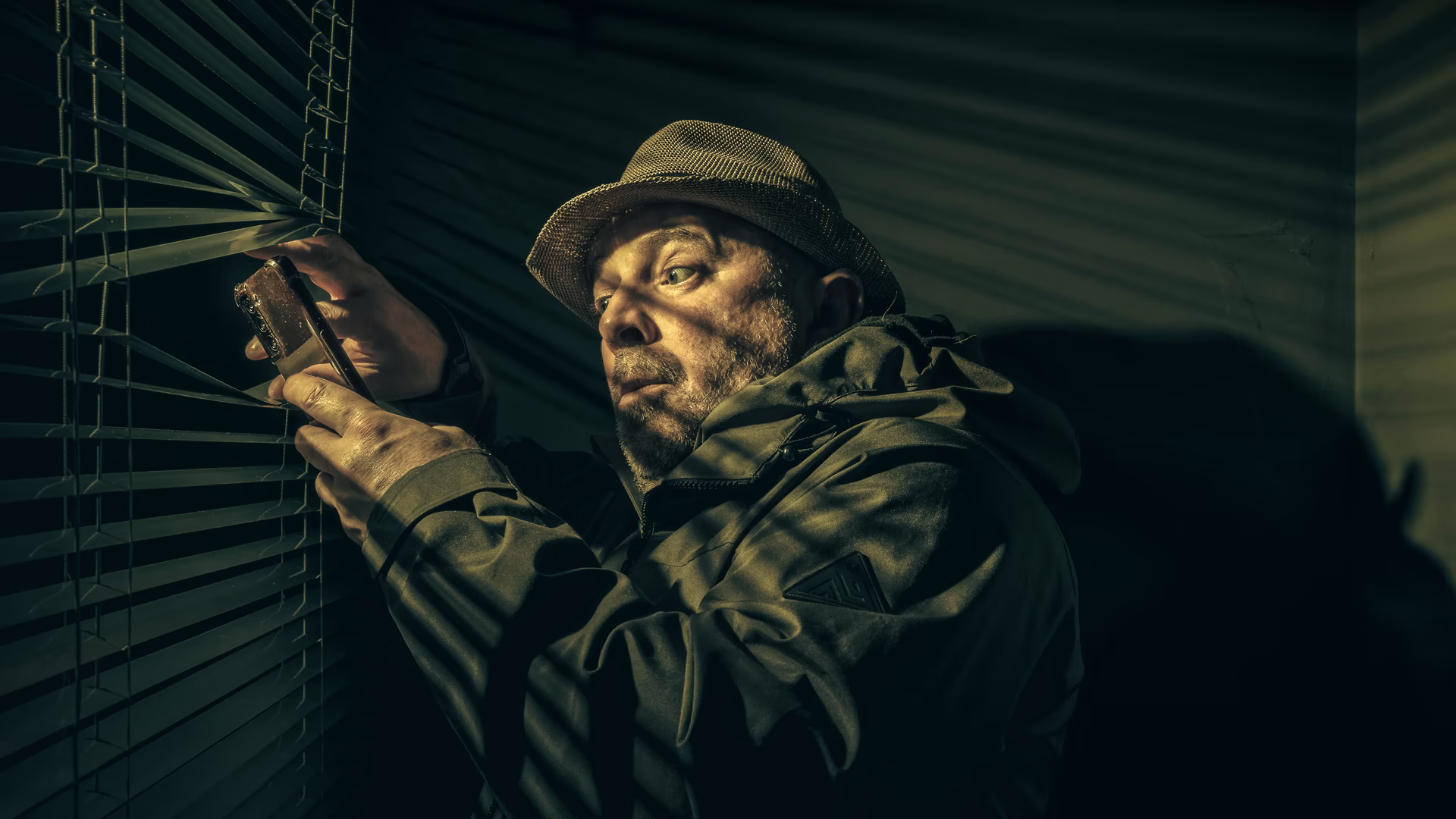 A man in a hat suspiciously peering through blinds, holding a phone in shadowy light - Image by Murry Lee from Pexels